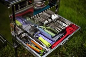 Fully stocked outdoor kitchen drawer with utensils, stored in our overland cooking kit, with grass in the background.