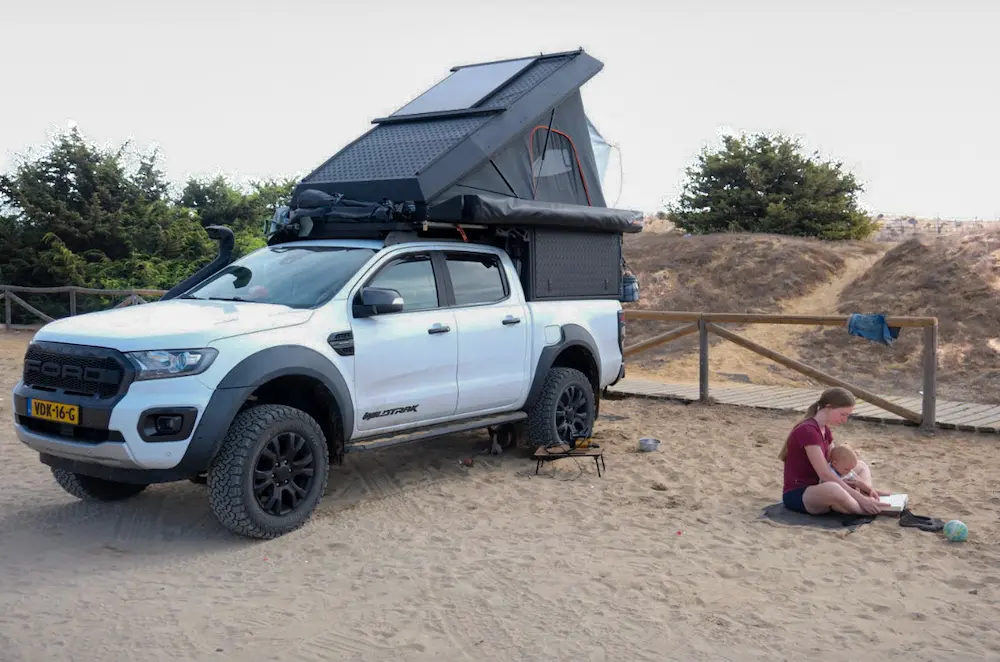 Mother reading a book with her toddler in her lap next to their 4x4 Ford Ranger with rooftop tent, while the dog rests under the car and a ball lies nearby. A folded table with a compressor is visible as they reinflate their tires after driving on sand.